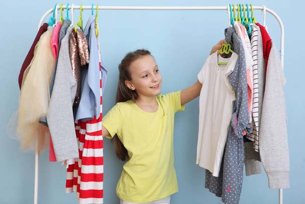 Χαριτωμένο χαρούμενο κοριτσάκι επιλέγει ρούχα με κρεμάστρες δαπέδου. Παιδικά ρούχα, παιδικά ψώνια. Μοντέρνα ρούχα για παιδιά. - Φωτογραφία, εικόνα