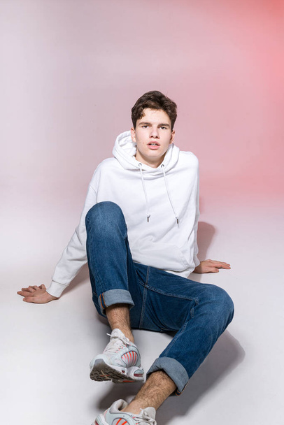 Young caucasian male model posing while sitting on the floor in the studio. Teenage boy in stylish clothes on a pink background. Jeans guy, white hood and sneakers. - Foto, Imagem