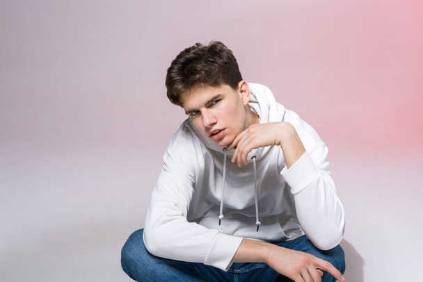 Handsome young stylish man sitting on the floor in studio. Teenager model posing while sitting on a pink background in casual clothes. The guy in jeans, sneakers and a white jacket with a hood. - Foto, imagen
