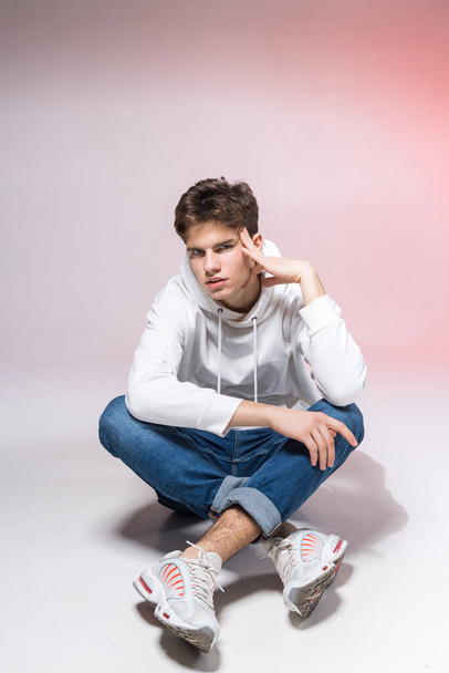 Mladý kavkazský mužský model pózující na podlaze ve studiu. Teenage chlapec ve stylovém oblečení na růžovém pozadí. Kluk v džínách, bílá kapuce a tenisky. - Fotografie, Obrázek