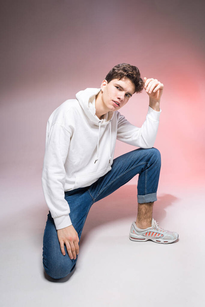 Handsome young stylish man sitting on the floor in studio. Teenager model posing while sitting on a pink background in casual clothes. The guy in jeans, sneakers and a white jacket with a hood. - Photo, Image