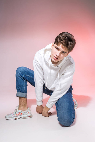 Handsome young stylish man sitting on the floor in studio. Teenager model posing while sitting on a pink background in casual clothes. The guy in jeans, sneakers and a white jacket with a hood. - Photo, Image