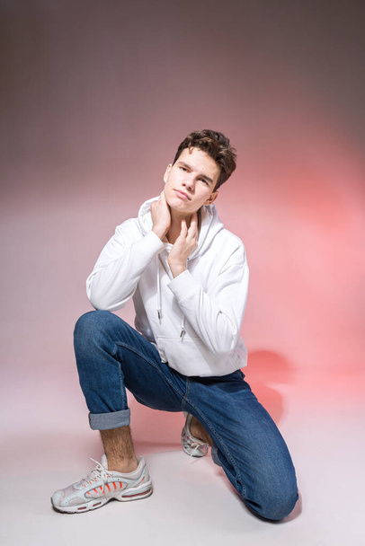Handsome young stylish man sitting on the floor in studio. Teenager model posing while sitting on a pink background in casual clothes. The guy in jeans, sneakers and a white jacket with a hood. - Zdjęcie, obraz