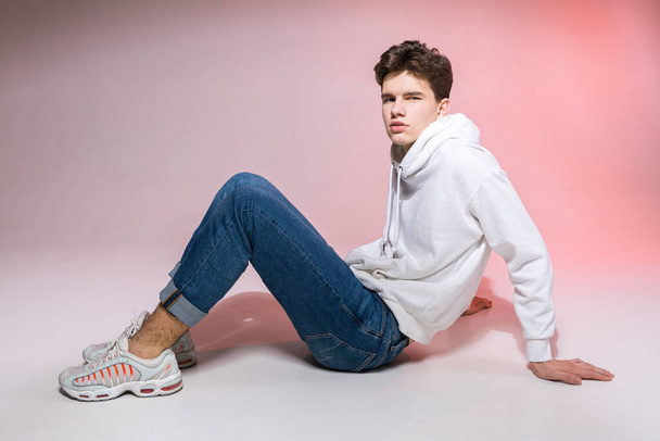 Pensive teen boy posing sitting on the floor in the studio. Portrait of stylishly dressed student. Studio shot Handsome attractive european man sitting on floor on a pink background. - Photo, Image