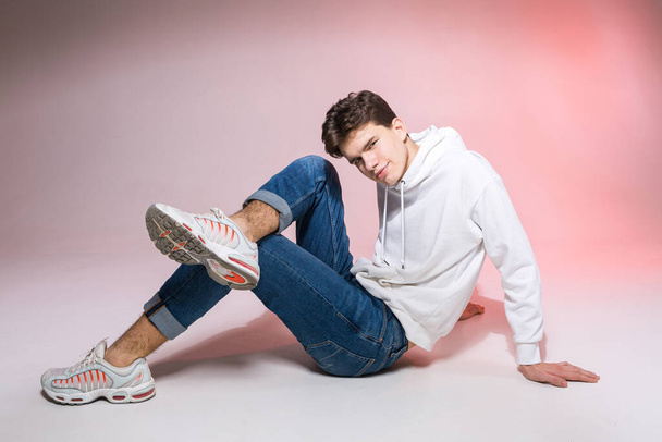 Handsome young stylish man sitting on the floor in studio. Teenager model posing while sitting on a pink background in casual clothes. The guy in jeans, sneakers and a white jacket with a hood. - Zdjęcie, obraz