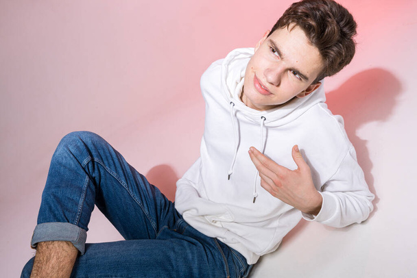 Modieus knap jong Europees mannelijk model gekleed in wit sweatshirt, blauwe jeans en witte sneakers poserend in studio op een roze achtergrond voor commerciële fotoshoot, kijkend naar camera. - Foto, afbeelding