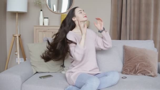 Smiling young woman playing with her long brunette hair sitting on sofa at home, slow motion - Záběry, video
