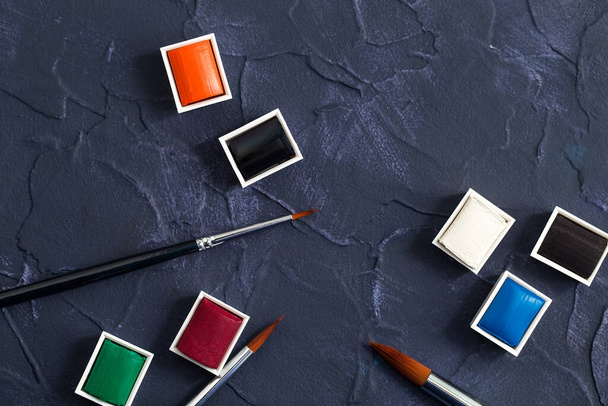 Aquarellfarben und Pinsel auf dunklem Hintergrund, Werkzeuge für eine kreative Werkstatt, Draufsicht - Foto, Bild