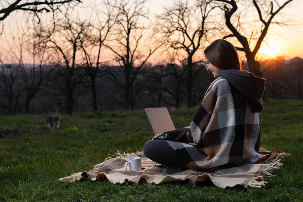 Hipster meisje ontwerper met een ruitje op haar schouders en bril met een laptop op haar schoot en een mok koffie ernaast, kijk op het laptopscherm - Foto, afbeelding