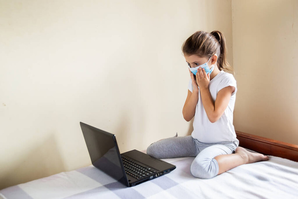 家庭用隔離コロナウイルスのパンデミック予防に滞在してください。小さな女の子はベッドの上に座ってラップトップを使用しています。予防流行。子供はテクノロジーを使う。COVIDによる遠隔オンライン教育- 2019. - 写真・画像