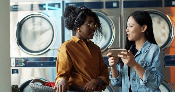 Multiethnic stylish young girls talking and watching photos or video on smartphone. Friends standing in laundry service. African American and Asian women with phone while washing machines working. - Fotó, kép
