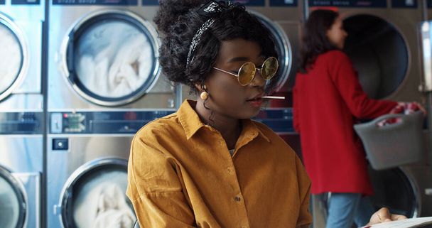 African American young beautiful girl in yellow glasses siting in laundry service room and flipping pages of journal. Woman with lollypop reading magazine while waiting for clothes to be washed. - Foto, afbeelding