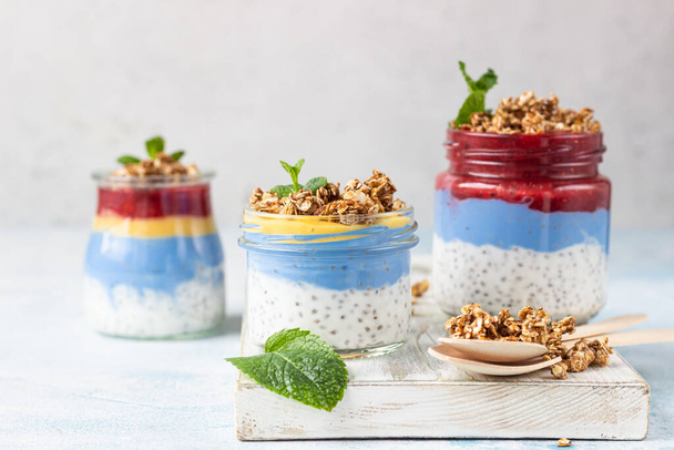 Vegan blue spirulina chia puddings with jam and granola in glass jars on a light blue table. Concept of healthy eating, dieting, fitness menu. Various layered dessert for breakfast.  - Φωτογραφία, εικόνα