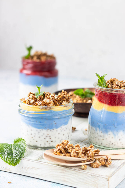 Vegan blue spirulina chia puddings with jam and granola in glass jars on a light blue table. Concept of healthy eating, dieting, fitness menu. Various layered dessert for breakfast.  - Foto, afbeelding