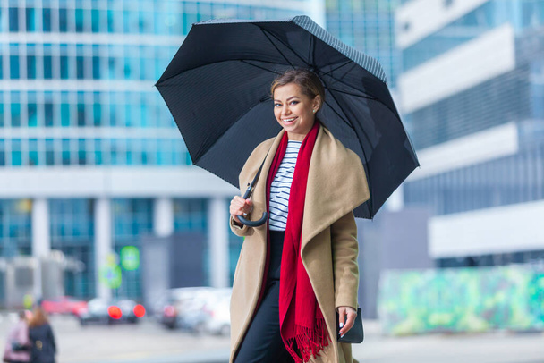 Rain. Outdoors lifestyle fashion portrait of stunning brunette girl. Walking on the city street. Going shopping. Wearing stylish white fitted coat, red neckscarf, black umbrella cane. Business woman. - Fotó, kép