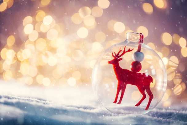 Christmas glass ball with a reindeer inside on snow. Lights glitter background - Photo, Image