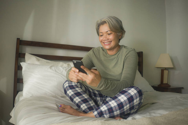 home lifestyle portrait of attractive and happy middle aged woman on her 50s using internet mobile phone in bed relaxed and cheerful online dating or enjoying social media app - Фото, изображение