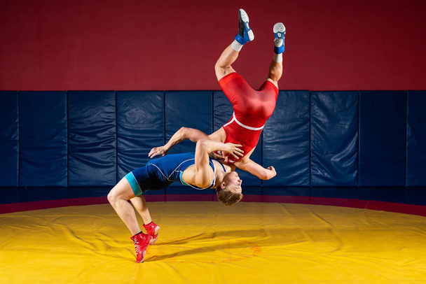 Dva silní muži v modrých a červených zápasnických punčocháčích zápasí a dělají suplexní wrestling na žlutém wrestlingovém koberci v tělocvičně. Zápasníci zápasí.  - Fotografie, Obrázek