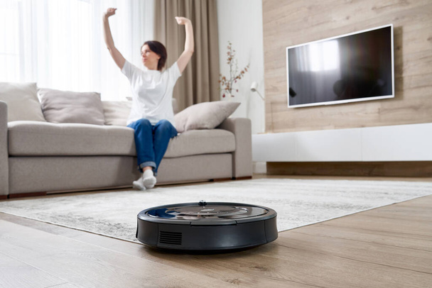 Robotic vacuum cleaner cleaning the room while woman resting on sofa - Фото, изображение