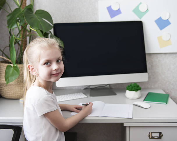 Залишайтеся вдома, щоб запобігти пандемії коронавірусу. Маленька дівчинка зі світлим волоссям сидить за столом і робить домашнє завдання
.  - Фото, зображення