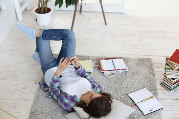Distance learning. Young female student studying at home, preparing for university exams, lying on floor against cozy domestic interior, surrounded with pile of books - Zdjęcie, obraz