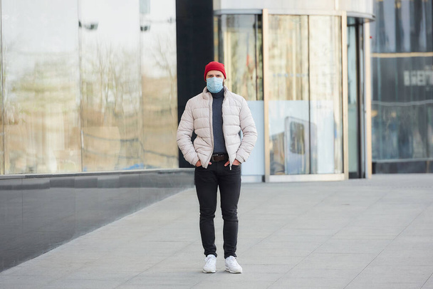 A man in a red watch cap wearing a medical face mask to avoid the spread coronavirus (COVID-19). A guy with a surgical mask on the face because of the pandemic walking dawn the street. - Fotografie, Obrázek
