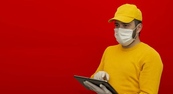 Sarı şapkalı teslimatçı, steril yüz maskesi eldivenli boş tişörtlü kırmızı arka planda izole edilmiş bir tablet tutuyor. Çalışan kurye servisi karantina virüsü koronavirüs konsepti. Dümdüz. Logo için yer - Fotoğraf, Görsel