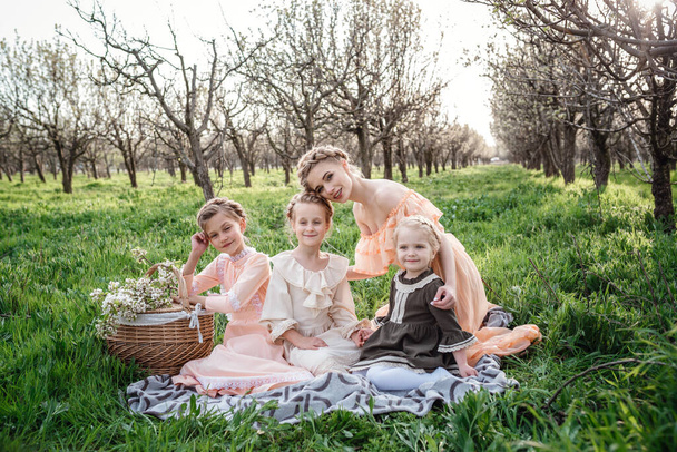 piękne dziewczyny i ich starsza siostra siedzi na kratę w ogrodzie z koszykiem pełnym kwiatów, ciesząc się wiosną. Idea i koncepcja przyjaznej rodziny i szczęśliwego dzieciństwa. Suknie retro - Zdjęcie, obraz