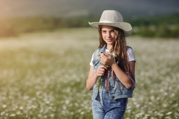 kalapban élvezi a tavaszt a százszorszép mezőn. A boldog gyermekkor koncepciója és eszméje. tinédzser farmerben overál a réten napsütéses nyári napon - Fotó, kép