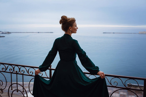 Όμορφη νεαρή γυναίκα περιμένει για μια εκδήλωση σε ένα εστιατόριο βεράντα. Προετοιμασία για τις γιορτές και τις εκδηλώσεις. Πολυτελές φόρεμα με θέα στη θάλασσα - Φωτογραφία, εικόνα