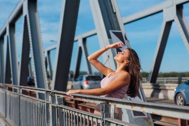 Warm summer portrait of a young beautiful woman posing on the old rusty transport bridge over the river during sunset. - Foto, Imagem