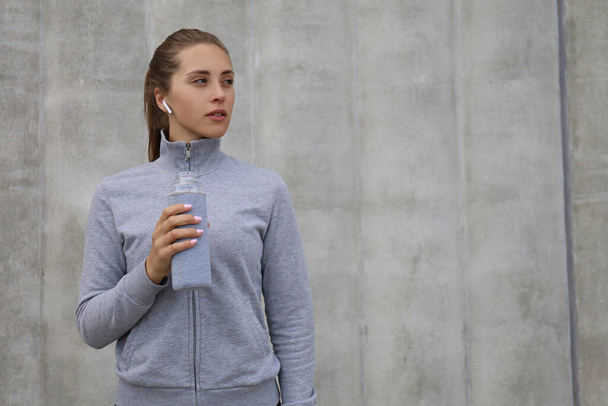 Beautiful female runner is standing outdoors holding water bottle. Fitness woman takes a break after running workout. - Foto, Imagem