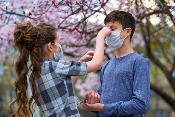 мальчик и девочка в маске на открытом воздухе, цветущие деревья, весенний сезон, время цветения - концепция аллергии и защиты здоровья от пыльного воздуха
 - Фото, изображение