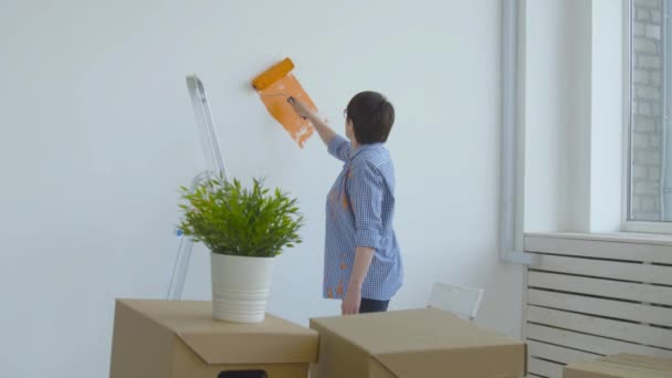 Flat renovation concept. Happy Middle-aged woman painting white wall with paint roller, orange paint - Footage, Video