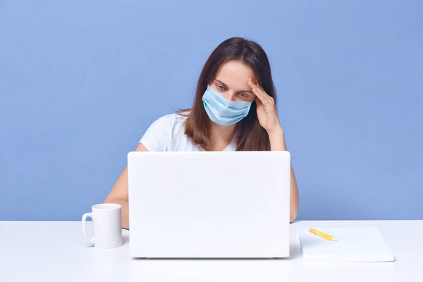 Caucasian woman wearing protective mask sitting in front of laptop. Protecting population from coronaviruses with protecting respiratory tract, girl working at home, looks tired, touching her head. - Foto, Imagem