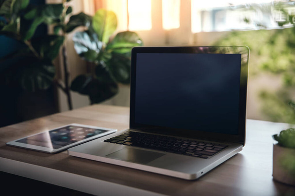 Laptop en tablet op houten bureau. Werken vanuit huis. Gezellige werkplek met raamlicht en groene plant - Foto, afbeelding