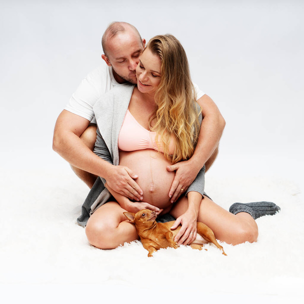 Любящий мужчина обнимает свою беременную жену сзади, сидя с милой собачкой на белом фоне. Материнство, беременность, новая жизнь и планирование семьи
 - Фото, изображение