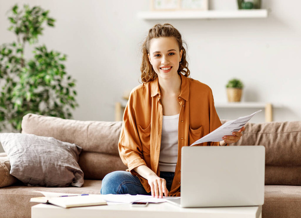 Vrolijke jonge vrouwelijke freelancer glimlach op de camera terwijl zitten op de zachte bank op de laptop en het controleren van gegevens in rapporten tegen wazig interieur van licht moderne apartmen - Foto, afbeelding