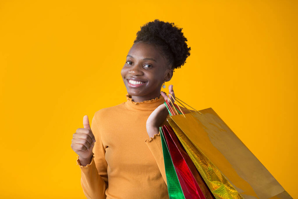 beautiful young african woman on yellow background with credit with packages in hand - Fotó, kép