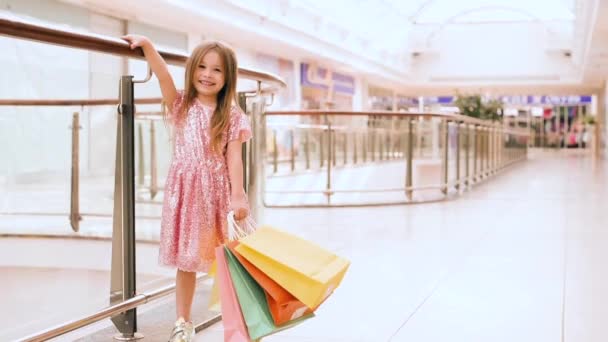 Portré egy boldog kislányról a plázában. Egy mosolygó, nevető lány rózsaszín ruhában, sokszínű táskákkal a kezében bevásárol. - Felvétel, videó