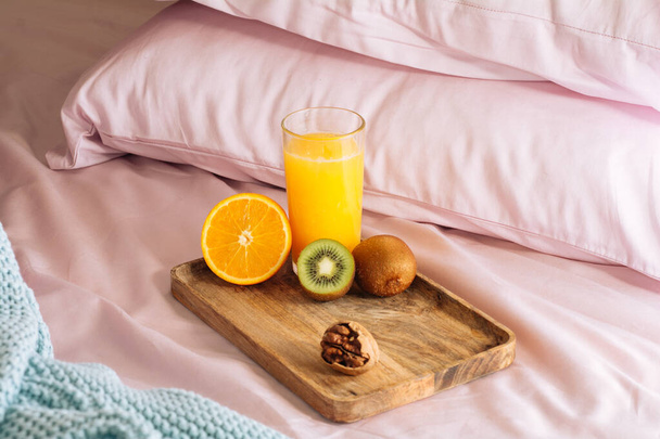 Morning healthy breakfast in bed with freshly squeezed orange juice, half orange, kiwi and walnut on a wooden tray against a backdrop of pink pillows and sheets and blue plaid - Fotó, kép