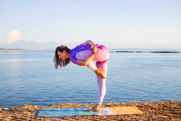 Aantrekkelijke vrouw die zich uitstrekt en zich voorbereidt op de yoga praktijk bij zonsopgang. Outdoor yoga praktijk in de buurt van de oceaan. Gezondheidszorg en zelfzorg concept. Op één been staan, in balans blijven. Bali, Indonesië - Foto, afbeelding