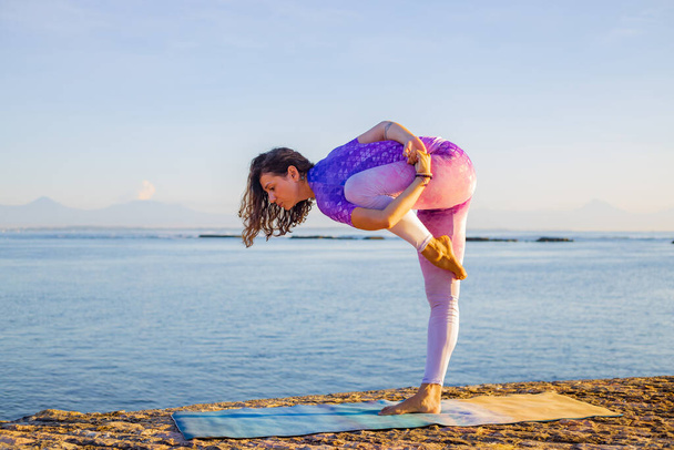 Mujer atractiva que se estira y se prepara para practicar yoga al amanecer. Practica yoga al aire libre cerca del océano. Concepto de salud y autocuidado. De pie en una pierna, mantener el equilibrio. Bali, Indonesia
 - Foto, Imagen