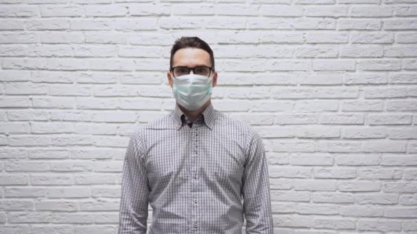 Φοβούμενος Καυκάσιος νεαρός επιχειρηματίας με καστανά μαλλιά με μαύρα γυαλιά σε καρό πουκάμισο με προστατευτική ιατρική μάσκα στο πρόσωπο απλώνει τα χέρια του προς τα πλάγια ενάντια σε λευκό τοίχο από τούβλα. Φοβάται.. - Πλάνα, βίντεο