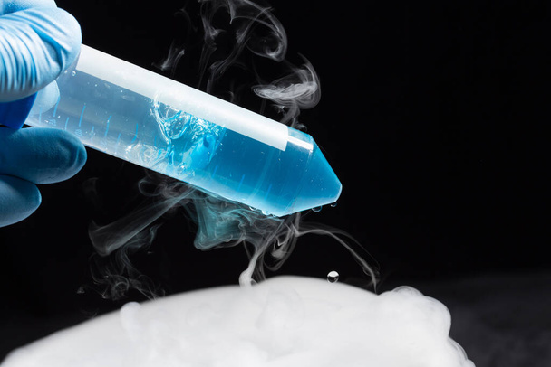Vloeibare stikstof stijgt tegen een zwarte achtergrond in een wetenschappelijk lab als farmaceutische werknemer voert tests uit op blauwe vloeistof in reageerbuis. - Foto, afbeelding