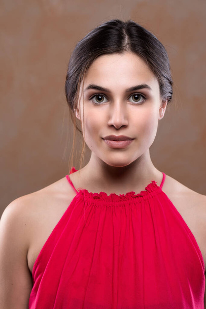 A confident young Caucasian girl in her mid-twenties poses for a portrait wearing a red blouse. Cosmetic make-up emphasizes the cheeks bones and plumps the lips. - Foto, Bild