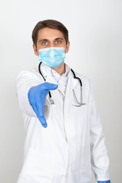 Handsome young physician wearing surgical mask is showing handshake on white background - Photo, Image