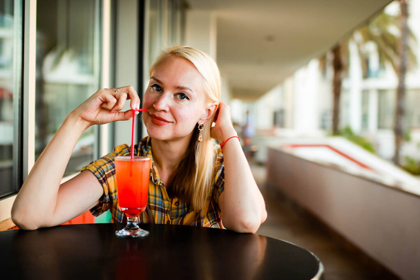 Portrét mladé blondýny popíjející koktejl a usmívající se, dívající se do kamery. - Fotografie, Obrázek