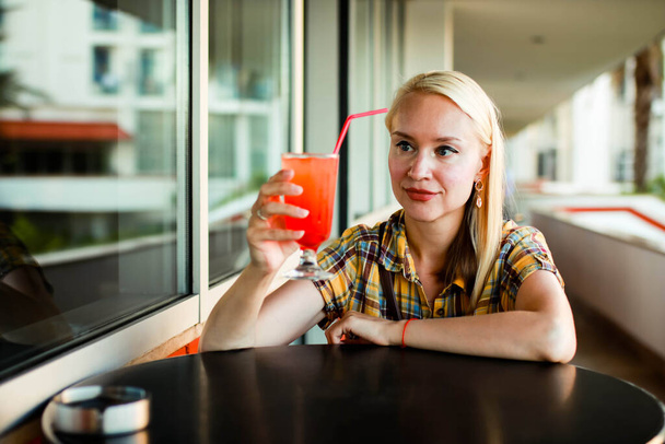 Mulher em uma camisa xadrez amarela está bebendo um coquetel de um copo em uma mesa em um restaurante
. - Foto, Imagem