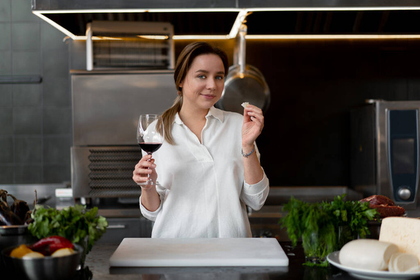 Beautiful young caucasian girl standing in kitchen in a white uniform smiling and tasting red wine Cute woman 30s years old in white shirt with food Ingredients cheese meat vegetables drinking - Foto, Bild